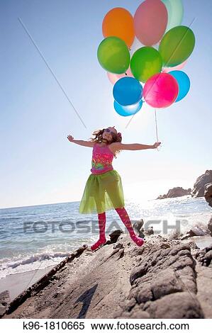 Stock Photography - little girl holding balloons and jumping while on the rocks at the ocean. Fotosearch