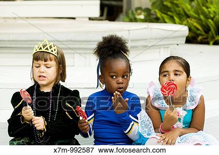 Stock Photo - Little girls Asian Indian, black & caucasian dressed in Halloween costumes enjoy their lollipops as one gets caught red-handed eating candy. Fotosearch