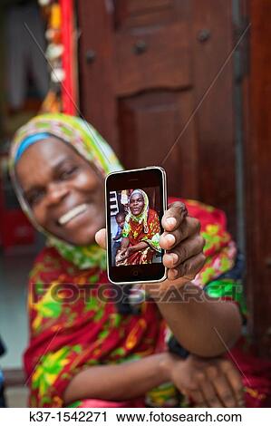 Stock Image - Local woman in East African market and holding smart phone with her photo.. Fotosearch