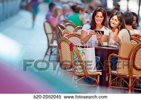 Stock Photography - Looking a smartphone. Women sisters 35 and 40 years taking coffee on a terrace. Donostia. San Sebastian. Gipuzkoa. Basque Country- Spain.. Fotosearch