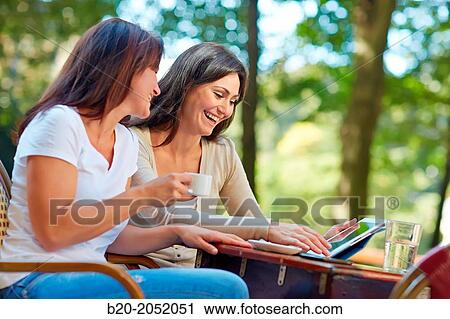 Stock Image - Looking a tablet. Women sisters 35 and 40 years taking coffee on a terrace. Donostia. San Sebastian. Gipuzkoa. Basque Country- Spain.. Fotosearch