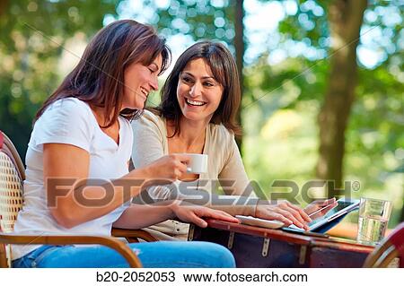 Stock Image - Looking a tablet. Women sisters 35 and 40 years taking coffee on a terrace. Donostia. San Sebastian. Gipuzkoa. Basque Country- Spain.. Fotosearch