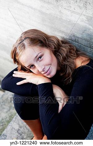 Stock Image - Lovely young woman sitting by a concrete wall looking at the camera. Fotosearch