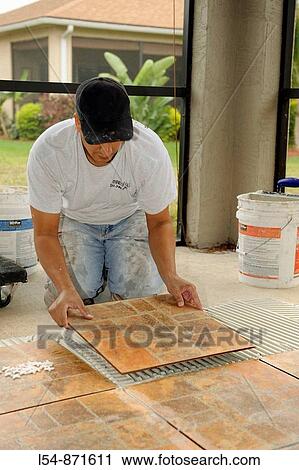 Stock Image - Male Hispanic Lays Ceramic Tile on Cement Floor in Patio Lanai. Fotosearch