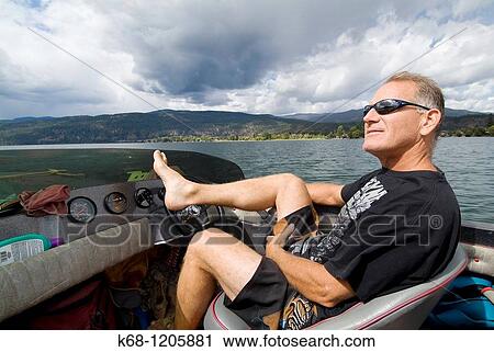 Stock Image - man, 50, drives a boat with his feet on Okanagan Lake, BC, Canada. Fotosearch