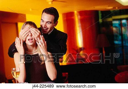 Stock Photo - Man covering eyes to a woman in a bar. Fotosearch