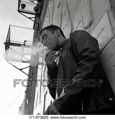 Stock Image - Man in alley, wearing a leather jacket and smoking a cigarette.. Fotosearch