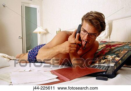 Stock Image - Man in boxers working and talking on cell phone in bed. Fotosearch