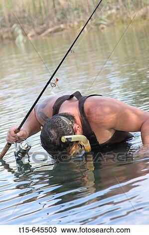Stock Image - Man in his 30's holding a fishing rod wearing snorkel mask looking for fish in the lake. Fotosearch