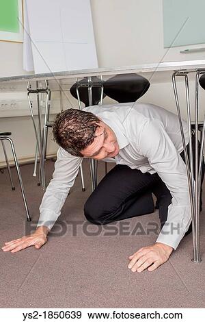 Stock Photo - Man in white shirt sitting under table in an office searching for something that is lost. Fotosearch