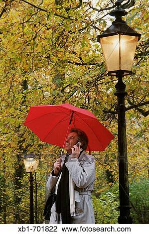 Stock Image - Man, middleaged, standing with an umbrella next to a street lamp, making a telephone call. Fotosearch