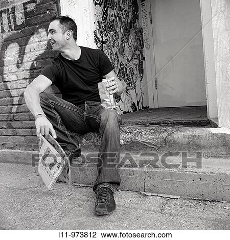 Stock Image - Man seated in alley, with newspaper and beer.. Fotosearch
