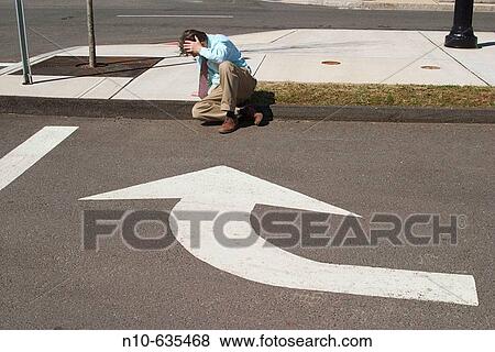 Stock Photo - Man sitting on a curb in front of a traffic arrow that points towards him. Fotosearch