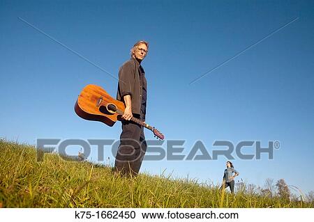 Stock Image - Man standing in field with guitar with woman coming over the hill. Fotosearch