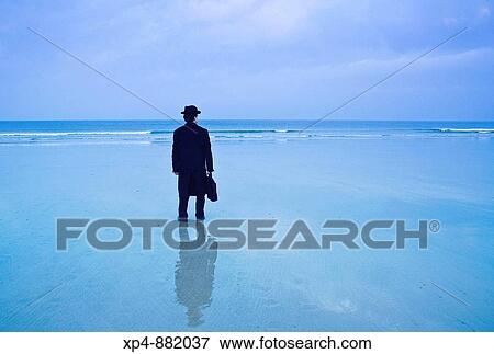 Man standing on the beach in black hat and overcoat with briefcase looks out to the ocean, his image reflected in the wet sand. View Large Photo Image Stock Photo - Man standing on the beach in black hat and overcoat with briefcase looks out to the ocean, his image reflected in the wet sand.. Fotosearch