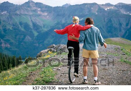 Stock Photograph - Man teaching woman how to ride a? monocycle? on top mountain. Colorado. USA. Fotosearch
