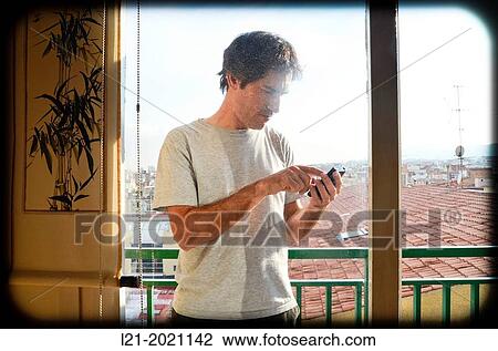 Stock Image - Man typing the phone on a balcony. In the background, a cityscape.. Fotosearch