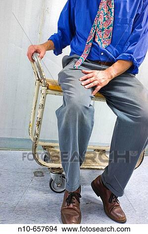 Picture - Man, wearing an uneven tie, sitting on a rolling cart.. Fotosearch