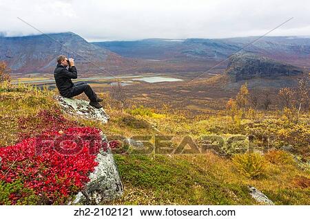Stock Image - Man with binoculars watching in to sarek national park and landscape are in autumn colors.. Fotosearch