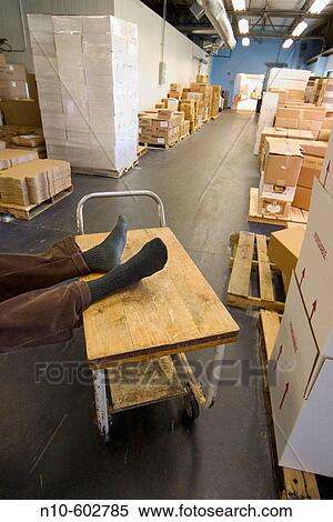 Stock Photography - Man's feet, wearing just socks, resting on a rolling cart in a factory's storage area.. Fotosearch