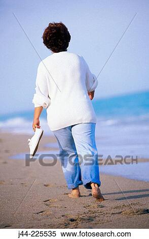 Stock Photography - Mature woman walking at the beach holding shoes in hand. Fotosearch