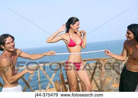Stock Photo - Men measuring woman's waist with bathing suit by the ocean. Fotosearch