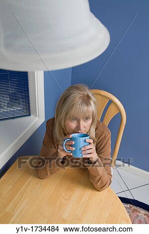 Picture - Middle-age blond woman drinking coffee in her kitchen. Fotosearch