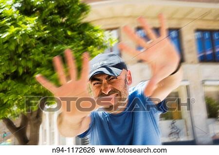 Stock Photograph - Middle-aged man with a beard and cap, in the center of the city, with open hands scold the photographer.. Fotosearch