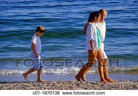 Stock Photo - Moody boy walking behind couple with pregnant woman on seashore. Fotosearch