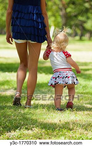 Stock Image - Mother holding hand of her female toddler while walking in a park. Fotosearch