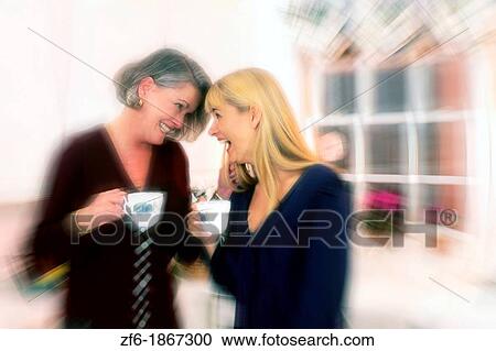 Stock Image - Mother in 50s and daughter in 20s having fun talking in kitchen while laughing and talking with coffee. Fotosearch