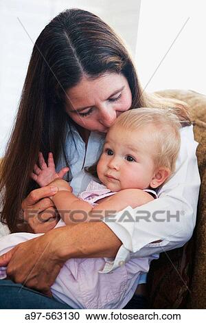 Stock Image - Mother kisses blonde infant girl on the head while holding her on her lap.. Fotosearch