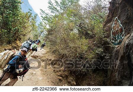 Stock Photograph - Nepal Himalayas Trekkers walking near Lotus flower symbol painted next to a mantra carved into the rockface and painted, outside the village of Bengkar Solukhumbu remote Mt Everest 55.. Fotosearch