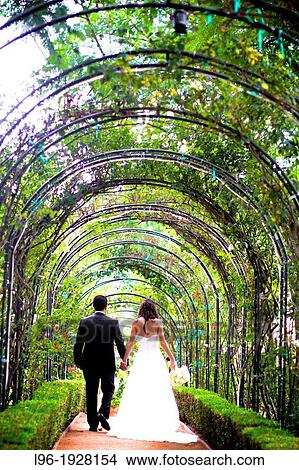 Picture - newly married couple walking hand in hand under a garden canopy arch away to their honeymoon. Fotosearch