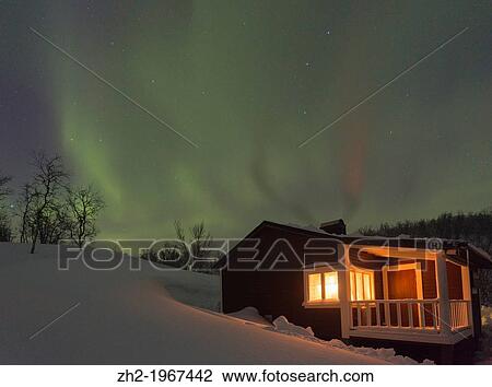 Stock Image - Northernlight, Aurora borealis, over a cottage with light coming through the window in winter season in march, in Riksgr? nsen, Swedish lapland, Sweden.. Fotosearch