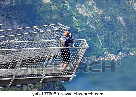 Stock Image - Obertraun, Salzkammergut, Austria, Europe Woman tourist looking through a Visio telescope on 5fingers viewing platform on Krippenstein mountain at the Dachstein World Heritage site with high view to Hallstattersee lake in the Austrian Alps. Fotosearch