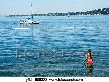 Stock Image - Paquis beach on Geneva Lake in Geneva, young woman going to swim in water of Geneva Lake, Lac Leman, summer vacation, sailboat in the background, Geneva, Switzerland. Fotosearch
