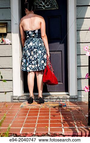Stock Photo - Partial back view of a woman wearing a dress, holding a red purse stading on a house porch in front of a door.. Fotosearch
