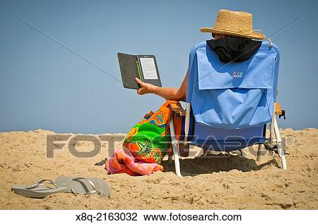 Stock Image - Person reading a book on a Kindle tablet device at the beach.. Fotosearch