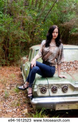 Stock Image - Portrait of a 20 year old brunettte woman sitting on an old car in a forest setting. Fotosearch