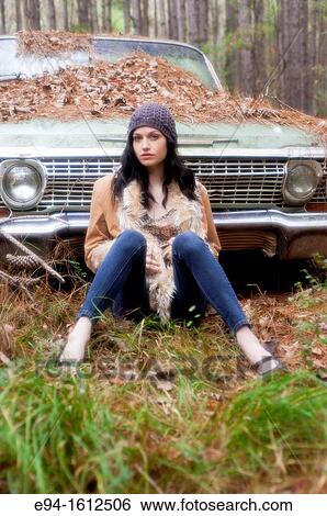 Portrait of a 20 year old brunettte woman wearing a knit hat and a leather coat sitting on the ground in front of an old car in a forest setting View Large Photo Image Stock Photograph - Portrait of a 20 year old brunettte woman wearing a knit hat and a leather coat sitting on the ground in front of an old car in a forest setting. Fotosearch