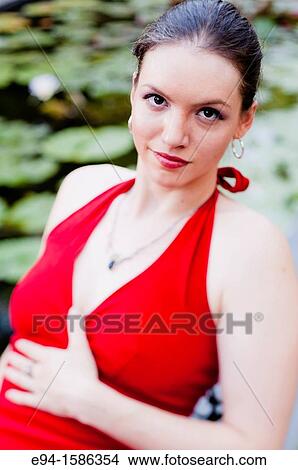 Picture - Portrait of a 24 year old brunette woman in a red dress sitting on a chair by a water lily pool looking at the camera. Fotosearch