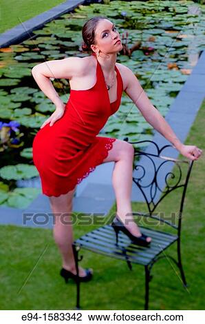Portrait of a 24 year old brunette woman in a red dress with one leg on a chair by a water lily pool looking at the camera View Large Photo Image Stock Image - Portrait of a 24 year old brunette woman in a red dress with one leg on a chair by a water lily pool looking at the camera. Fotosearch
