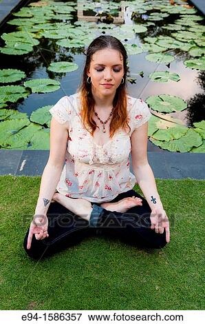 Stock Photo - Portrait of a 24 year old brunette woman sitting in the lotus postion on grass with water liliies in the background. Fotosearch