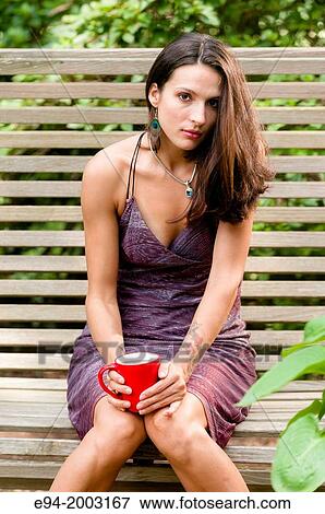 Stock Photo - Portrait of a 27 year old brunette woman in a summer dress sitting on a bench looking at the camera in an outdoor setting holding a cup of coffee.. Fotosearch