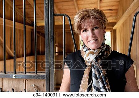 Stock Image - Portrait of a 52 year old blonde woman in casual black dress in a horse barn setting looking at the camera.. Fotosearch