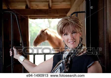 Portrait of a 52 year old blonde woman in casual black dress in a horse barn setting looking at the camera with a horse in the background. View Large Photo Image Stock Image - Portrait of a 52 year old blonde woman in casual black dress in a horse barn setting looking at the camera with a horse in the background.. Fotosearch