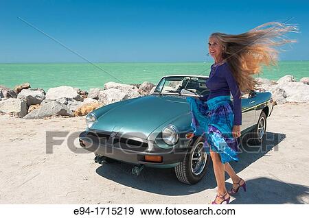 Stock Photo - Portrait of a 57 year old woman standing by her MG car at a beach. Fotosearch