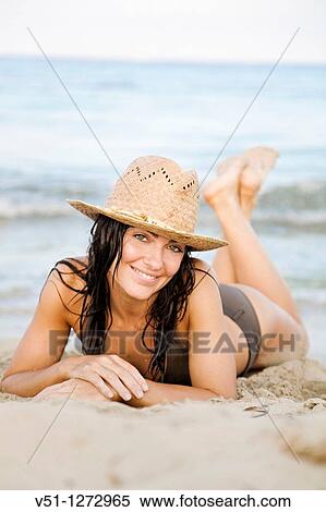 Stock Photography - Portrait of a beautiful brunette woman smiling a camera at the beach with bikini and a hat. Fotosearch