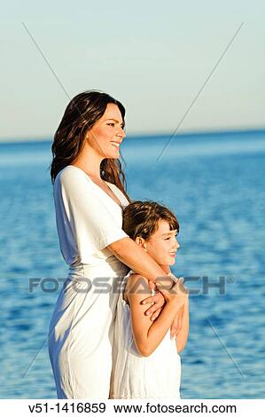 Stock Photo - Portrait of a cute woman looking at horizon with daughter at the beach. Fotosearch
